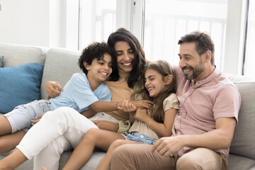 Cheerful parents embracing tickling little kids on home couch, enjoying active games, leisure, family activities with children, laughing, having fun with giggling son and daughter