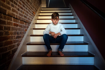 Sad and lonely barefoot boy in white sweater sitting alone on a staircase