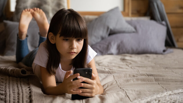 Overusing phone. Little school age girl lie on bed with cell focused on screen has internet addiction dependency on gadgets. Preteen child spend much time online feel information overload. Copy space