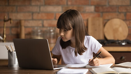 Homeschooling online. Focused little latin girl primary school pupil engaged in distant learning do homework write exercise. Preteen child study from home watch lesson on laptop take notes at kitchen