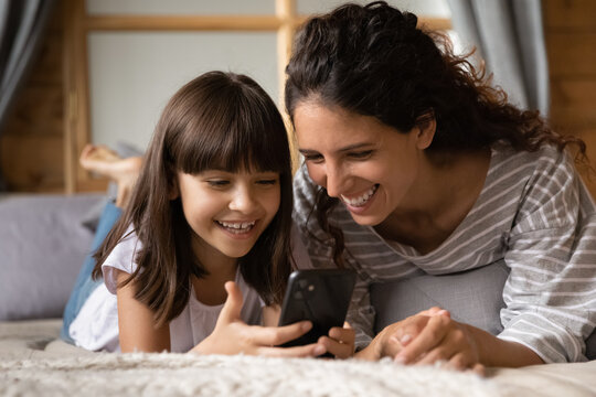 Pleasant leisure with cell. Excited young latin female mom or elder sister spend time lying on bed with cute little girl daughter using smartphone app. Mum and child enjoy pastime with phone together