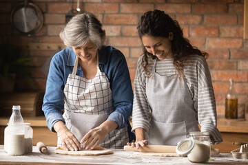 Doing cookies at home. Friendly older latin mother and daughter in law in kitchen aprons kneading rolling dough on table talk enjoy cooking together. Caring grownup child assist aged mom in baking pie - obrazy, fototapety, plakaty