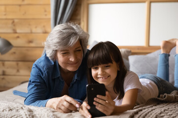 Happy little latin girl lie on bed close to old age grandmother show new photo video at social network account. Joyful preteen grandchild make cute selfie on cell webcam together with senior granny