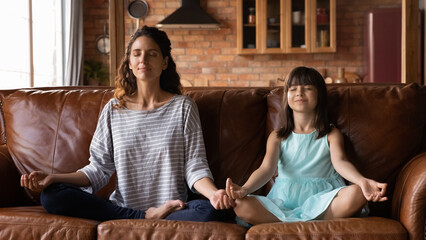Mom and daughter yogis. Calm hispanic mother and little girl practice yoga exercise sit on sofa in...