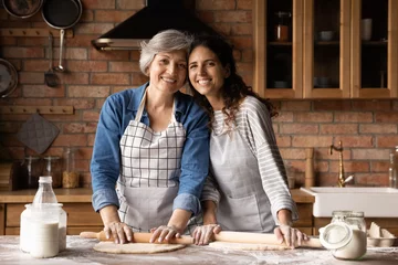 Foto op Aluminium Portrait of two cheerful latina women retired mother young adult daughter wear aprons hug at kitchen table look at camera distracted of rolling dough. Senior granny grown grandkid cook homemade bakery © fizkes