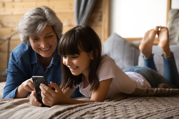 Happy preteen kid granddaughter rest on large bed with loving retired hispanic granny buying toys on ecommerce web site play cellphone game in online app. Mature babysitter use phone with little girl - obrazy, fototapety, plakaty