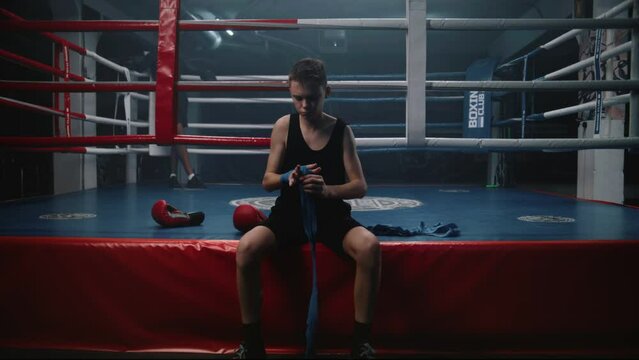Young boxer sits near boxing ring and wraps his hands with bandage for martial arts. Teenage athlete prepares to fight tournament or competition, trains in boxing gym. Physical activity. Dolly shot.