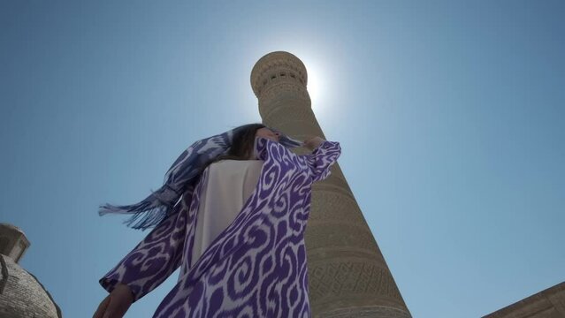 A young woman in a white dress and a long oriental robe dances in the sunshine next to an ancient minaret