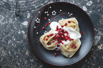 Black plate with steamed dumplings manty, pomegranate seeds and yogurt, horizontal shot on a dark-brown granite surface, high angle view