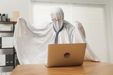 Overworked ghost. Man covered with white sheet using laptop at wooden table in office