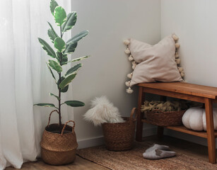 A cozy corner of the living room - a wooden bench with a pillow and a blanket, baskets, a ficus flower, slippers on a jute carpet. A cozy house