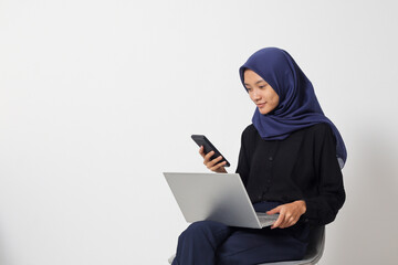 Portrait of excited Asian hijab woman in casual shirt sitting on chair, working on laptop while using a mobile phone. Businesswoman concept. Isolated image on white background