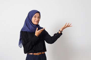 Portrait of excited Asian hijab woman in casual shirt promoting product, pointing finger to the side. Advertising concept. Isolated image on white background