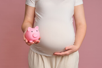 Pregnant woman holding piggy bank of money, studio pink background. The concept of pregnancy and...