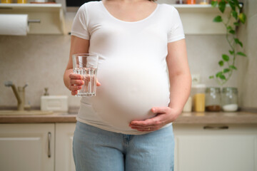 A pregnant woman holds a glass of clean water in her hands, home kitchen background. Pregnancy and...