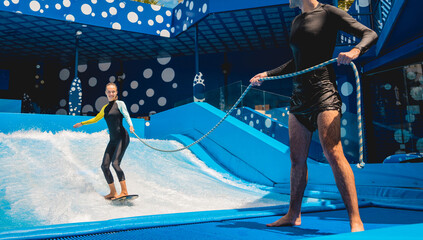 Beautiful young woman surfing with trainer on a wave simulator at a water amusement park