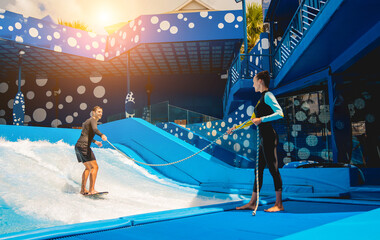 Young man surfing with trainer on a wave simulator at a water amusement park
