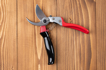 Steel gardening secateurs, scissors tool with red and black grip for pruned of plants and flowers garden work, on a wooden table background. Open state. Top view.