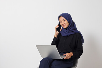 Portrait of excited Asian hijab woman in casual shirt sitting on chair, working on laptop while having conversation on smartphone call. Businesswoman concept. Isolated image on white background