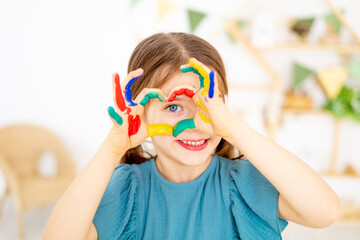 Naklejka premium cute little baby girl with colorful painted stained hands in finger paints shows a heart in the nursery and learns to draw or fooling around, art lessons with pencils and paints, close-up