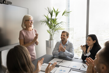 Satisfied diverse team of employees applauding mature female boss, leader on meeting, laughing,...