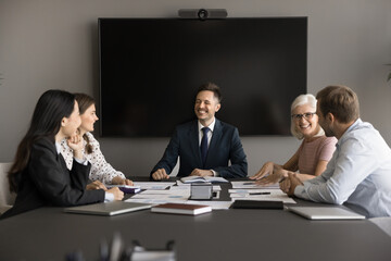 Cheerful young business team leader man holding teamwork meeting with colleagues. Diverse colleagues talking at large table in boardroom, brainstorming, laughing, having fun