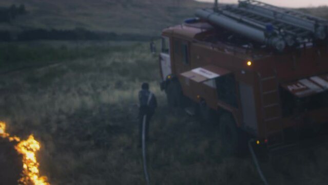 A real Firefighter extinguishes a burning flame from a hose with water. Nature is on fire. Water from a hose is sprayed onto the fire. There is a fire engine nearby