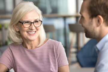 Happy attractive senior businesswoman talking to younger colleague man on business meeting, smiling, laughing, enjoying networking, discussing cooperation, teamwork, partnership