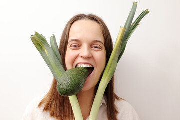Crazy Caucasian woman holding green leek and avocado isolated over gray background keeps diet...