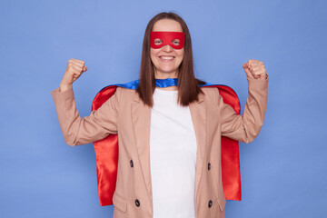 Strong joyful attractive brown haired woman wearing superhero costume posing isolated over blue background standing with raised arms showing her strength