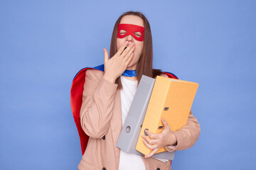 Tired sleepy overworked brown haired woman wearing superhero costume holding documents in folders...