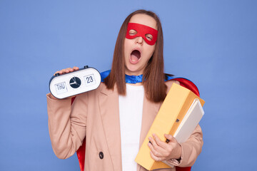 Sleepy tired brown haired woman wearing superhero costume holding documents in folders posing isolated over blue background showing alarm clock yawning with widely open mouth