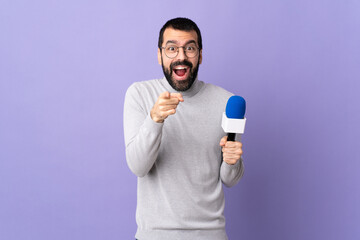 Adult reporter man with beard holding a microphone over isolated purple background surprised and...