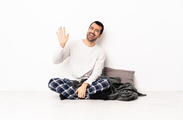 Caucasian man in pajamas sitting on the floor at indoors saluting with hand with happy expression