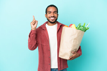 Young latin man holding a grocery shopping bag isolated on blue background pointing up a great idea