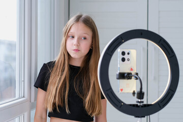 Beautiful teenage girl posing in front of a phone on a tripod. Child blogger shoots video in front of camera and ring lamp