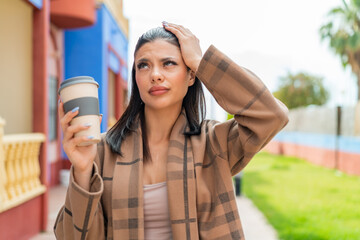 Young pretty woman holding a take away coffee at outdoors having doubts and with confuse face expression