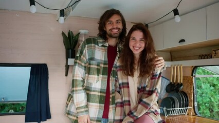 Portrait of a happy couple, a brunette guy with stubble in a green checkered shirt hugs his brunette girlfriend in a pink checkered shirt in their campervan outside the city in a camp during a picnic