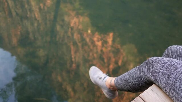 woman's legs moving on river water while she is sitting on a bridge, reflection of stone cliff on the surface