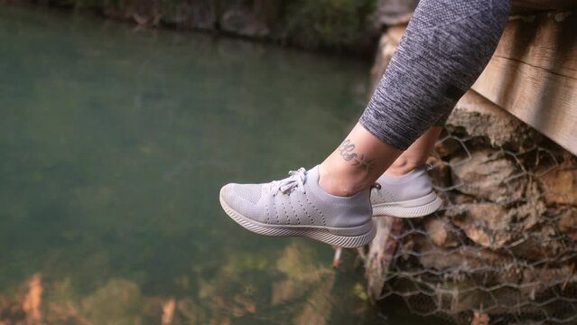 Woman's feet moving on river water while she is sitting on a bridge, reflection of stone cliff on the surface