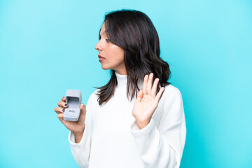 Young hispanic woman holding a engagement ring isolated on blue background making stop gesture and...