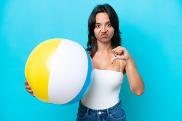 Young hispanic woman holding beach ball isolated on blue background showing thumb down with...