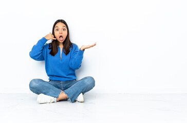Young mixed race woman sitting on the floor isolated on white background making phone gesture and doubting