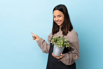 Young brunette mixed race woman holding a plant over isolated blue background pointing back.