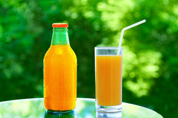 Orange juice in a glass bottle and glass on a mirror tabletop in the summer outdoors against a background of green foliage.