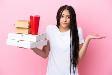 Teenager girl with braids holding pizzas and burgers over isolated pink background having doubts...