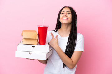 Teenager girl with braids holding pizzas and burgers over isolated pink background pointing to the...
