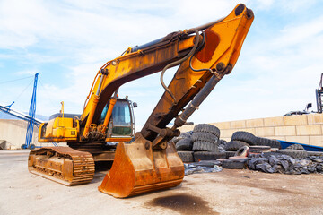 A crawler excavator stands on the port grounds next to a pile of old tires.