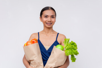 A cheerful young Asian woman holds paper bags full of fresh fruits and vegetables on a white...