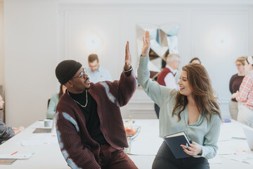 Two cheerful colleagues celebrate a successful team effort with a high-five in a lively office setting, efficiently capturing the essence of teamwork and job satisfaction amongst diverse co-workers.